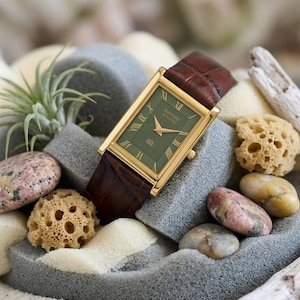 Puede incluir: Un reloj rectangular Seiko con caja dorada, esfera verde oscuro con &iacute;ndices de n&uacute;meros romanos y correa de cuero marr&oacute;n con patr&oacute;n de cocodrilo texturizado. La esfera del reloj muestra manecillas doradas y un peque&ntilde;o logotipo.