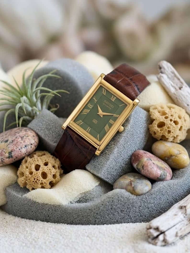 Puede incluir: Un reloj rectangular Seiko con caja dorada, esfera verde oscuro con &iacute;ndices de n&uacute;meros romanos y correa de cuero marr&oacute;n con patr&oacute;n de cocodrilo texturizado. La esfera del reloj muestra manecillas doradas y un peque&ntilde;o logotipo.