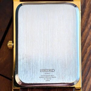Puede incluir: Un reloj de pulsera rectangular en tono dorado con una correa de cuero marr&oacute;n. La parte posterior del reloj presenta una placa de acero inoxidable cepillado con el texto SEIKO, QUARTZ, WATER RESISTANT, STAINLESS STEEL BACK, 60226714 y JAPAN.