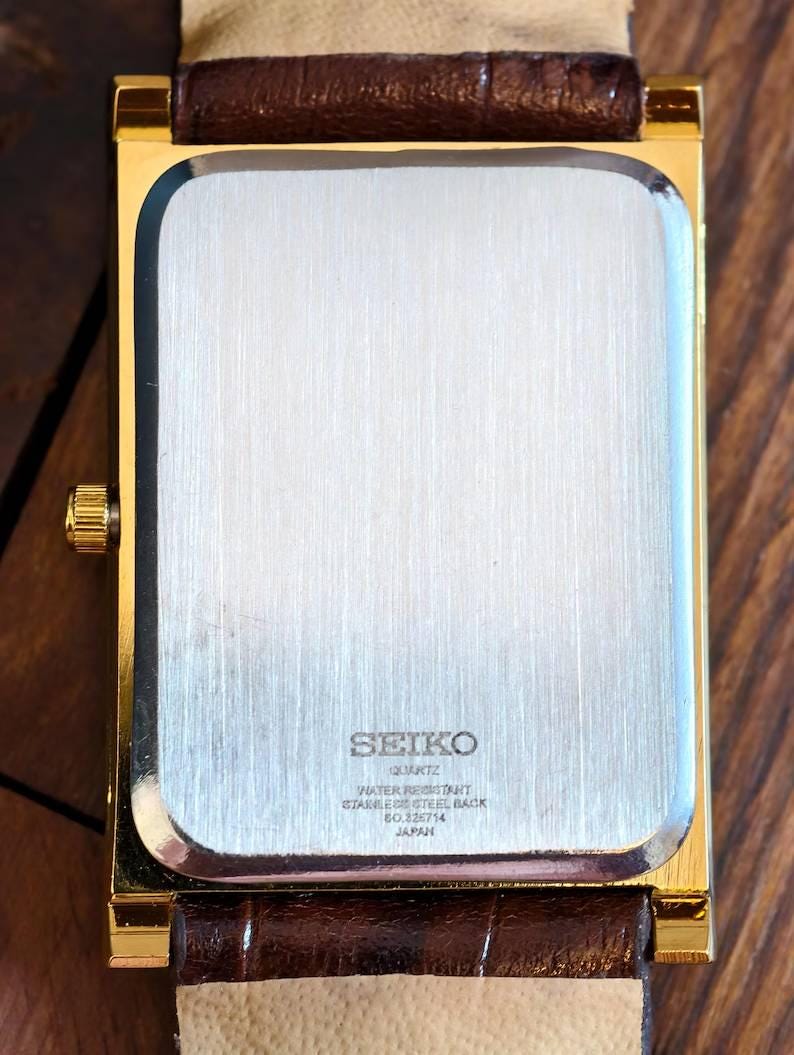 Puede incluir: Un reloj de pulsera rectangular en tono dorado con una correa de cuero marr&oacute;n. La parte posterior del reloj presenta una placa de acero inoxidable cepillado con el texto SEIKO, QUARTZ, WATER RESISTANT, STAINLESS STEEL BACK, 60226714 y JAPAN.