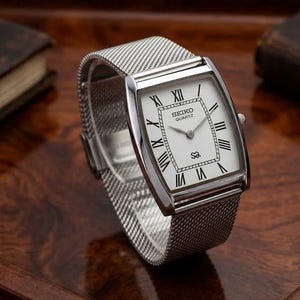 Könnte beinhalten: Eine silberfarbene Seiko Quartz Armbanduhr mit rechteckigem Gehäuse, weißem Zifferblatt, schwarzen römischen Ziffern als Stundenmarkierungen und einem feinen Metall-Mesh-Armband. Das Zifferblatt zeigt das Seiko Quartz Logo und ein SQ-Symbol.