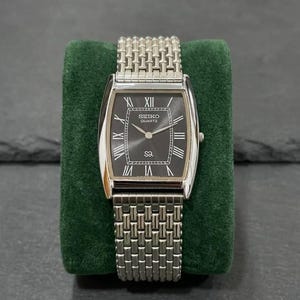 May include: A rectangular Seiko Quartz watch with a silver-tone case and a dark gray dial. The watch features Roman numeral hour markers and a silver-tone bracelet. The watch is displayed on a green velvet surface.