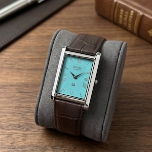 May include: A rectangular Seiko Quartz watch with a silver case and a turquoise face. The watch has Roman numeral hour markers and a brown leather band with a crocodile pattern. The watch is displayed on a gray velvet watch pillow.