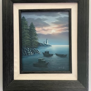 May include: Framed landscape painting featuring a coastal scene with a lighthouse, boats, and trees. The artwork is set against a twilight sky with a full moon. The painting is enclosed in a black wooden frame with a white inner border.
