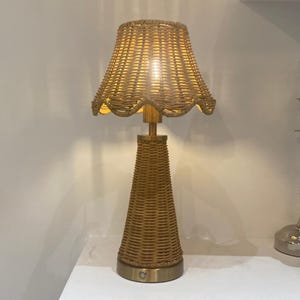 May include: A decorative table lamp with a woven rattan shade and base. The shade has a scalloped edge, and the lamp has a brass-colored metal stem and base. The lamp is lit, emitting a warm glow. The lamp is a home decor item.