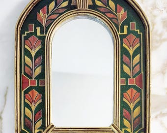 Pode incluir: Um espelho de parede arqueado com uma moldura dourada e uma borda decorativa sobre um fundo verde escuro. A borda é adornada com padrões florais repetitivos em tons de laranja, amarelo e vermelho profundo, criando um design simétrico.