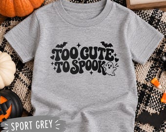 May include: A sport grey short-sleeve t-shirt featuring black text that reads "TOO CUTE TO SPOOK" in a retro font, accompanied by graphic illustrations of a small ghost, two bats, and several four-pointed stars scattered around the lettering.