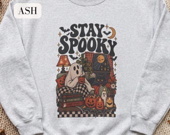May include: An ash gray crewneck sweatshirt featuring a graphic print with the text STAY SPOOKY. The illustration depicts a ghost sitting on a couch with a black cat, surrounded by pumpkins, candles, books, and bats in a cozy room setting.