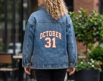 May include: A blue denim jacket featuring the text OCTOBER 31 in orange block letters on the back, paired with dark gray jeans and gray sneakers. The jacket has a classic collar, long sleeves, and a button-up front, worn over a simple outfit.