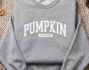 May include: A heather grey long-sleeve sweatshirt featuring the white text PUMPKIN SEASON printed in a bold, arched collegiate font across the chest. The garment has a crew neckline, ribbed cuffs, and a ribbed hem, showcasing a simple design.