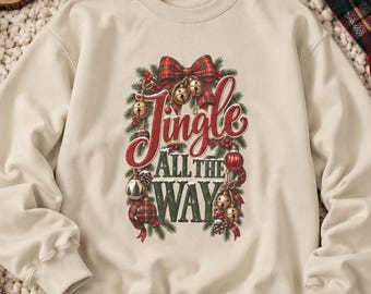 May include: A beige crewneck sweatshirt featuring a central graphic with the text Jingle All The Way in red and dark green lettering. The design includes illustrations of pine branches, red and gold ornaments, and a red plaid bow.