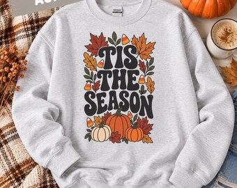 May include: An ash gray crewneck sweatshirt featuring a graphic print with the text 'TIS THE SEASON' in black, surrounded by colorful autumn leaves, orange pumpkins, and candy corn illustrations on the front center of the garment.