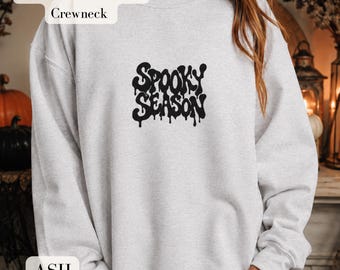 May include: An ash gray crewneck sweatshirt featuring the text "SPOOKY SEASON" printed in a black, dripping, stylized font across the center chest. The garment is labeled as a Gildan 18000 unisex crewneck, showcasing long sleeves and a ribbed collar.