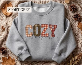 May include: A sport grey Gildan crewneck sweatshirt featuring the word COZY in a multi-colored plaid pattern. The letters are filled with various shades of brown, green, and red tartan, with the word season written in a black cursive script below.