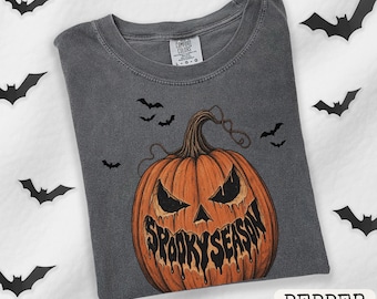May include: A charcoal gray short-sleeve t-shirt featuring a large orange jack-o'-lantern graphic with a dripping black text design that reads "SPOOKY SEASON" across the mouth. The shirt is decorated with small black bat silhouettes.