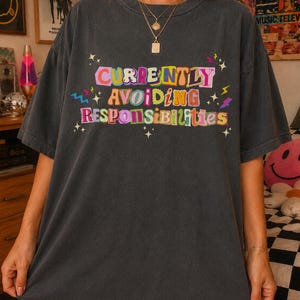 May include: A charcoal gray short-sleeve t-shirt featuring the colorful text "CURRENTLY AVOIDING RESPONSIBILITIES" in a playful, multi-font style. The design is accented with small star and lightning bolt graphics in yellow, pink, and blue.