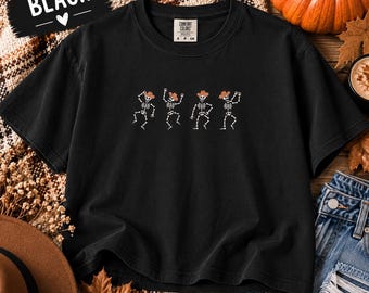 May include: A black short-sleeve t-shirt featuring a graphic of four white skeletons wearing brown cowboy hats in various dancing poses. The shirt has a crew neckline and a Comfort Colors brand label on the inside of the collar.