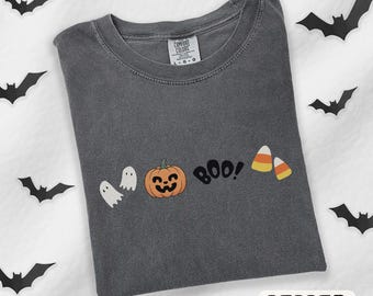 May include: A charcoal gray crew neck t-shirt featuring a graphic print of two white ghosts, an orange jack-o'-lantern, the black text "BOO!", and two pieces of candy corn. The shirt has a Comfort Colors label at the inner neckline.