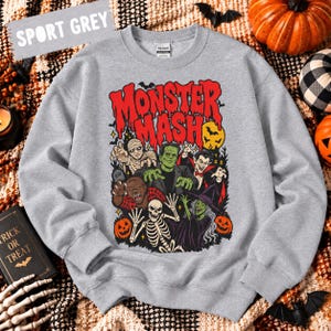 May include: A sport grey crewneck sweatshirt featuring a graphic print with the text "MONSTER MASH" in red. The illustration depicts a mummy, Frankenstein's monster, a vampire, a werewolf, a skeleton, and a witch surrounded by pumpkins.