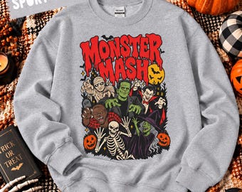 May include: A sport grey crewneck sweatshirt featuring a graphic print with the text "MONSTER MASH" in red. The illustration depicts a mummy, Frankenstein's monster, a vampire, a werewolf, a skeleton, and a witch surrounded by pumpkins.