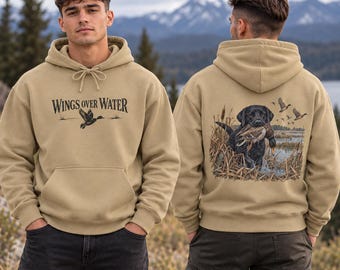 May include: A tan hooded sweatshirt featuring the text WINGS OVER WATER and a flying duck on the front. The back displays a graphic of a black Labrador retriever carrying a duck in its mouth amidst reeds, with flying ducks in the sky.
