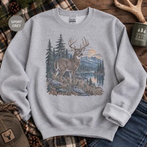 May include: A light gray crewneck sweatshirt featuring a detailed graphic print of a deer standing in a forest landscape with mountains, a lake, and a sunset. The sweatshirt has long sleeves, a ribbed collar, and a Gildan brand label at the neck.