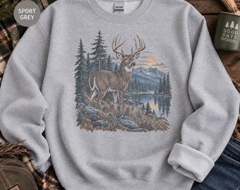 May include: A light gray crewneck sweatshirt featuring a detailed graphic print of a deer standing in a forest landscape with mountains, a lake, and a sunset. The sweatshirt has long sleeves, a ribbed collar, and a Gildan brand label at the neck.