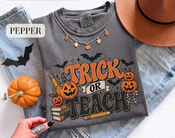 May include: A charcoal gray t-shirt featuring a graphic print with the text TRICK OR TEACH, orange pumpkins, stacked books, pencils, and black bats. The shirt is displayed with blue denim jeans, a brown hat, and a necklace with small charms.