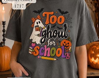 May include: A charcoal gray t-shirt featuring a graphic print of a ghost wearing a polka dot bow, holding books and a coffee cup. The text reads Too ghoul for school, surrounded by bats, a spider web, a pumpkin, pencils, and school supplies.