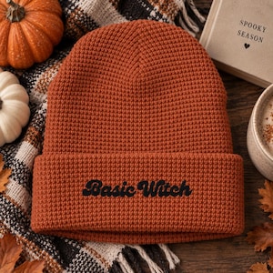 May include: A burnt orange waffle-knit beanie with a folded cuff. The text "Basic Witch" is embroidered in black thread on the front of the cuff. The hat features a textured grid pattern throughout the fabric and a rounded crown construction.