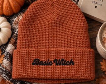 May include: A burnt orange waffle-knit beanie with a folded cuff. The text "Basic Witch" is embroidered in black thread on the front of the cuff. The hat features a textured grid pattern throughout the fabric and a rounded crown construction.
