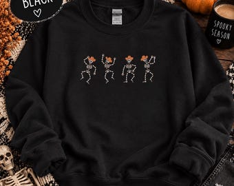 May include: A black long-sleeve crewneck sweatshirt featuring a graphic print of four white skeletons wearing orange cowboy hats in various dancing poses. The garment has a ribbed collar, cuffs, and hem, with a Gildan brand label at the neckline.
