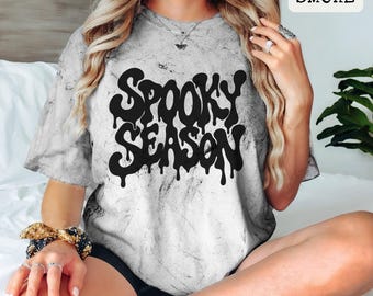 May include: A short-sleeve t-shirt with a gray and white acid-wash pattern. The front features the black text "SPOOKY SEASON" in a stylized, dripping font. A small label in the upper right corner displays the text "SMOKE" in black letters.