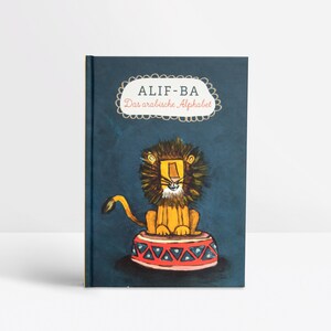 Könnte beinhalten: Ein Kinderbuch mit marineblauem Einband. Es zeigt einen Cartoon-Löwen auf einer rot-weißen Zirkusplattform. Der Buchtitel "ALIF-BA Das arabische Alphabet" wird in einem weißen, dekorativen Banner angezeigt.