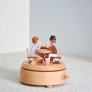 Op de afbeelding: Een houten muziekdoos met een barscène met twee figuren die aan een tafel zitten en elk een roze beker vasthouden. Er is ook een kleine hond aanwezig. De doos is lichtbruin met een slinger en kleine pootjes.