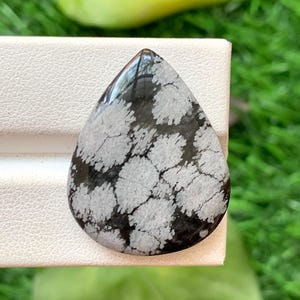 May include: A teardrop-shaped snowflake obsidian gemstone featuring a polished, smooth surface with a distinct black base colour and contrasting white, radial, flower-like patterns scattered across the stone's face in a natural, organic arrangement.