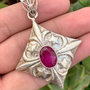 May include: A diamond-shaped silver pendant featuring a central oval-cut deep pink gemstone. The silver surface is textured with raised star motifs and decorative scrollwork, topped with an intricate filigree bail for a chain attachment.