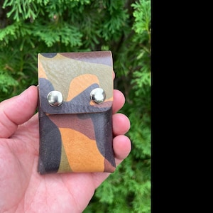May include: A small rectangular pouch featuring a camouflage pattern in shades of brown, tan, olive green, and black. The pouch has a fold-over flap secured by two silver-toned metal snap buttons on the front surface, constructed from leather.