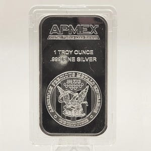 May include: A silver bar encased in a clear plastic holder. The bar is marked with the text "APMEX", "1 TROY OUNCE .999 FINE SILVER", and an image of an eagle. The bar is displayed on a clear plastic stand.