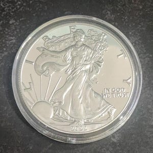 May include: A silver coin with a detailed depiction of a woman in flowing robes, rays of sunlight, and the inscription "IN GOD WE TRUST." The coin is encased in a clear plastic holder. The year "2022" is visible on the coin.