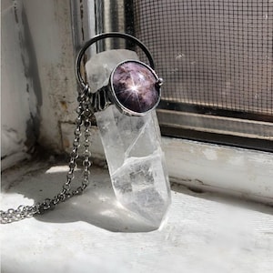 Puede incluir: Un collar con colgante de plata con un cristal de cuarzo transparente. El colgante tiene un engaste circular con una piedra oscura púrpura en forma de estrella. El collar se exhibe en un alféizar blanco. En el fondo, una mosquitera.