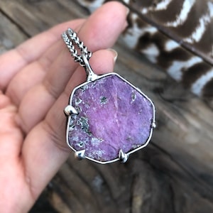 May include: A silver pendant featuring a large, irregular-shaped, purple gemstone. The stone has a rough, textured surface with hints of green. The pendant is held by a silver chain and is displayed against a wooden background.