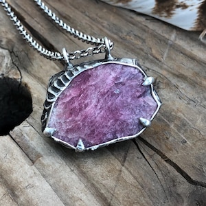 Può includere: Una collana d'argento con un grande pendente irregolare in pietra preziosa rosa. Il pendente è incastonato in argento e pende da una delicata catena. La gemma ha una superficie strutturata.