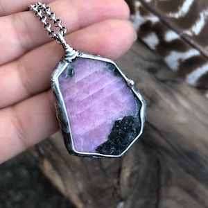 May include: A silver-toned pendant necklace featuring a large, irregularly shaped gemstone. The gemstone is primarily a light pink color with a dark green and black section. The pendant is attached to a delicate chain.