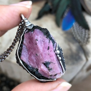 May include: A pendant featuring a raw, jagged pink tourmaline crystal embedded in a dark, textured matrix. The stone is encased in a dark, oxidized silver metal setting with small decorative spheres, suspended from a dark metal chain link necklace.