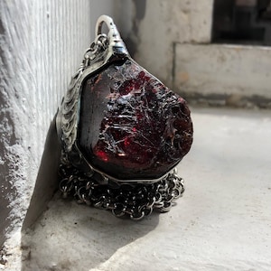 Op de afbeelding: Een ruwe, dieprode granaat hanger met een zilverkleurige metalen setting en ketting. De edelsteen is onregelmatig van vorm met een getextureerd oppervlak. De hanger wordt gepresenteerd op een wit oppervlak, wat de natuurlijke schoonheid van de steen benadrukt.