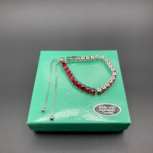 May include: A silver-toned bracelet with red square-cut stones rests on a green box. The bracelet has a sliding clasp and is made with Swarovski crystal. The box has a white sticker that says "Made with Swarovski Crystal."