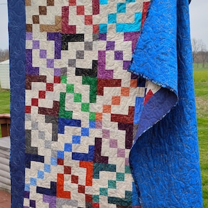 Puede incluir: Un edredón hecho a mano con un diseño de patchwork con cuadrados en varios colores, incluyendo rojo, azul, verde y morado. El edredón tiene un borde azul oscuro y un reverso azul liso. El edredón se sostiene, mostrando su diseño.