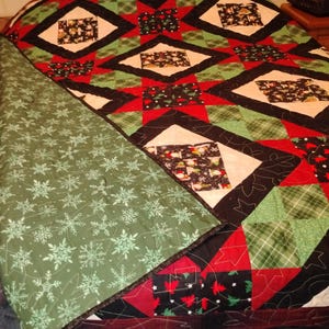 May include: A quilted blanket featuring a patchwork design of red, green, and black fabric squares with holiday-themed prints. The top side displays diamond-shaped patterns, while the folded corner reveals a green fabric with white snowflake motifs.
