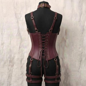 May include: A dark burgundy corset featuring a high neck collar with a buckle, adjustable shoulder straps, and a lace-up back closure. The garment includes attached garter straps with metal buckles that wrap around the upper thighs.